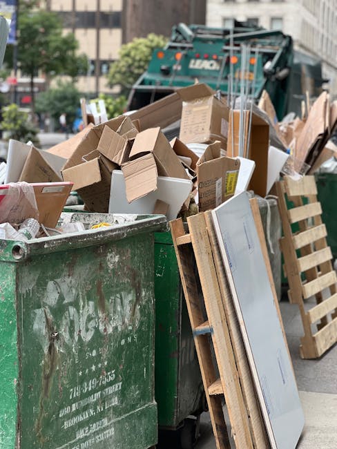 A large green waste bin filled with an assortment of cardboard boxes, some flattened and others still in regular shape, along with a white plastic container and various packing materials. The cardboard varies in size and color, ranging from light brown to darker shades, and some have printed labels or tape. Leaning against the bin is a wooden pallet with evenly spaced slats, possibly used for transporting bulk items. In the background, a green waste collection truck is partially visible, with its rear section equipped for waste loading, situated on a paved street or driveway. Surrounding the scene are trees and nearby buildings, indicating an urban or residential environment. The lighting appears natural, suggesting daytime conditions. The scene depicts an instance of private waste disposal, aligning with independent rubbish clearance services provided by Waste Clearance Swiss Cottage, as part of their rubbish removal offerings in the local area.