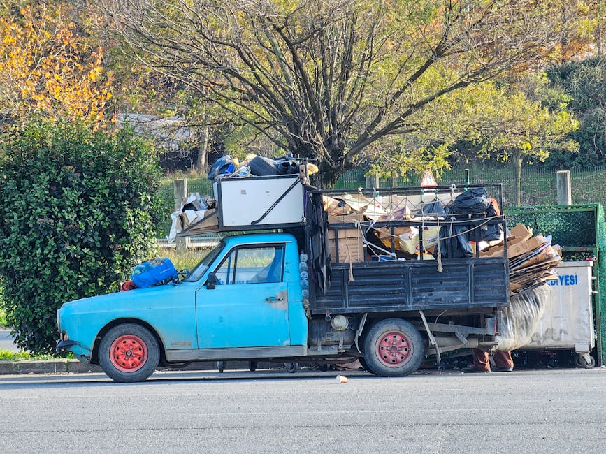 A compact blue pickup truck with a slightly weathered finish and red wheels parked on the side of a street; the truck's cargo bed is loaded with various types of waste, including flattened cardboard boxes, black plastic bags, and miscellaneous debris, which are secured with ropes and partially extending over the sides. The cargo also includes a large white appliance or container, possibly an old microwave or oven, situated at the front of the load, with reflective surfaces and a slight metallic sheen. Behind the truck, a neatly trimmed green hedge and a large deciduous tree with a mixture of green and yellow leaves are visible, indicating a park or residential area setting. The environment appears well-lit with natural daylight, and the scene suggests an instance of private waste disposal or a local rubbish removal service, as part of an independent collection process supported by Waste Clearance Swiss Cottage. The street surface is asphalt, contributing to a typical urban or suburban environment suitable for rubbish clearance activities.
