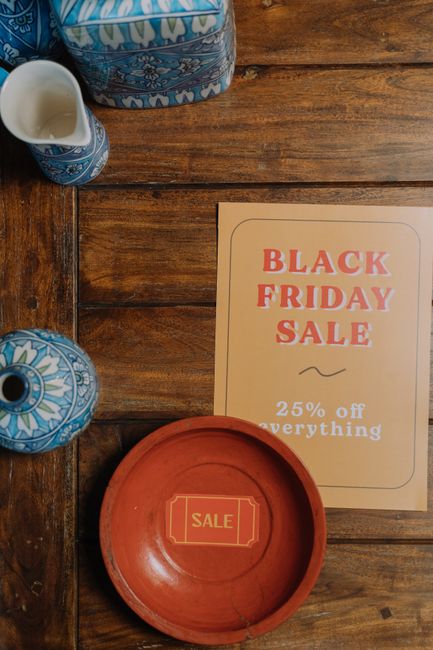 A wooden surface with a promotional flyer for a Black Friday sale, advertising 25% off everything, positioned next to a small decorative blue and white ceramic vase and a matching blue and white ceramic pitcher, both featuring intricate patterns. Below these items, there is a round terracotta-colored bowl with the word 'SALE' inscribed on its interior. The scene suggests a retail or market setting, with the flyer indicating a special discount event, and the ceramics adding a decorative touch that could be found in a boutique or store prepared for promotional displays. The background consists of the natural wood grain of the surface, creating a warm, rustic environment. The overall scene is well-lit, emphasizing the textures of the ceramics and the printed flyer, with a neutral tone that aligns with themes of urban clearance or retail surroundings where items are packed away or cleared during promotional events. Waste Clearance Swiss Cottage's service relevance may relate to managing excess merchandise or organizing clearance stock in retail premises.