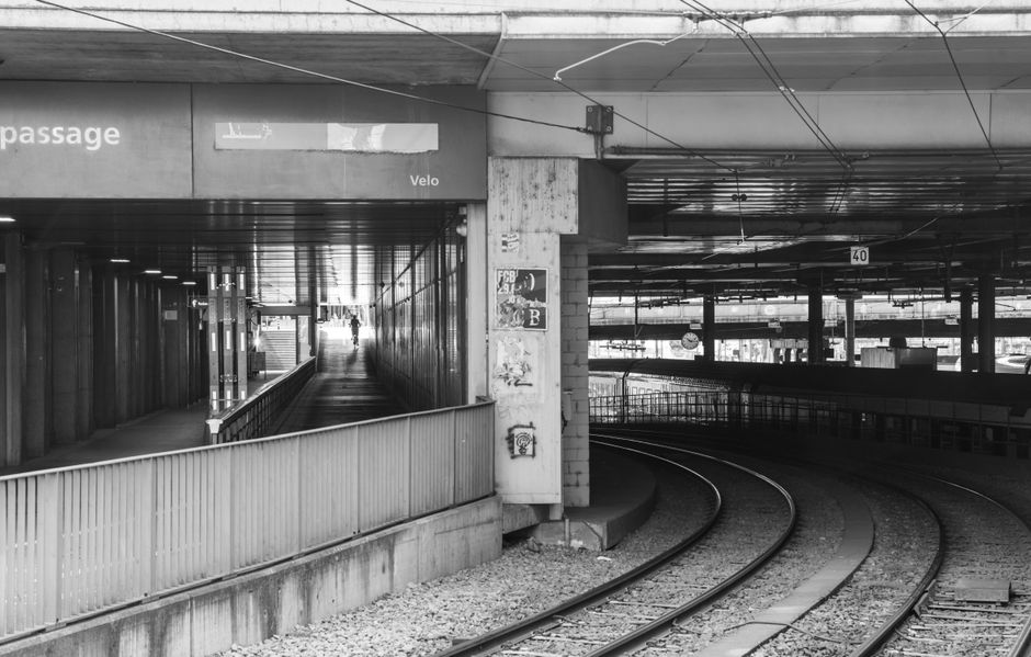 The image depicts an urban train station underpass with a curved railway track running through the foreground, constructed of concrete and metal, with visible gravel and ballast surrounding the rails. On the left side, there is a narrow, descending pedestrian walkway enclosed by metal railings, leading towards an illuminated exit that appears to open to the outside environment. The walls of the underpass are made of concrete and feature some weathered surfaces with peeling paint and small posters adhered to the wall on the right. Overhead, a network of electrical wires and support fixtures run parallel to the ceiling, which is composed of concrete slabs with visible joints. The lighting is dim, predominantly illuminated by artificial light sources, creating a subdued atmosphere. This scene relates to the context of waste clearance for urban infrastructure, as Waste Clearance Swiss Cottage provides services that could include clearing debris and waste from such transport facilities during maintenance or private site clearance, ensuring safe and efficient operation of public transport environments.
