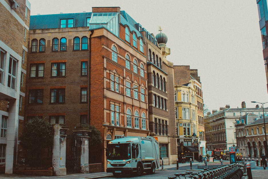A street scene featuring a blue waste collection truck parked along the curb in front of a row of historic multi-storey buildings with a mix of brick and ornate architectural detailing. The truck, positioned in the foreground, is part of a rubbish removal service and has a clean, metallic finish with a cab area above the compacted waste area. Behind the truck, traditional-style buildings with large bay windows, decorative moldings, and pitched roofs are visible, some with small domed turrets or ornamental features. To the right, a pedestrian pathway is lined with bollards, with additional historic and modern buildings extending into the distance. The environment appears urban with minimal traffic, subdued lighting indicating overcast weather, and no visible pedestrians. This scene subtly reflects alternative waste handling methods, such as private rubbish collection, with Waste Clearance Swiss Cottage operating in the local area to facilitate rubbish removal in such urban settings.